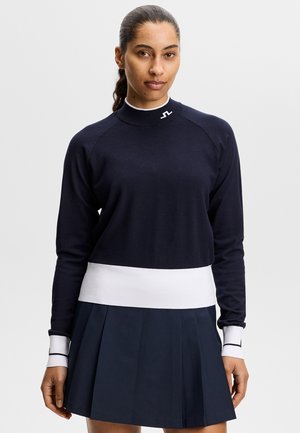 Frau mit zurückgebundenen dunklen Haaren, die einen marineblauen Pullover mit weißen Manschetten und Saum sowie einen marineblauen plissierten Rock trägt und vor einem schlichten Hintergrund steht.