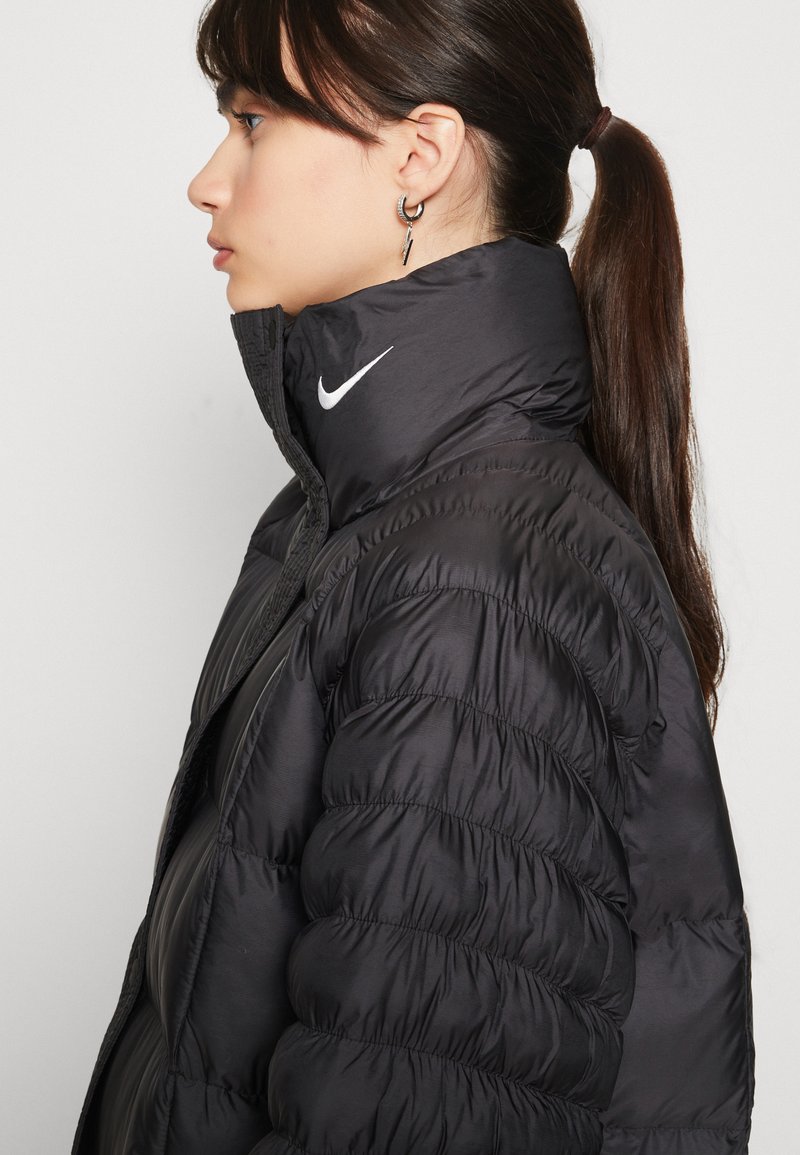 Czarna puchowa kurtka z wysokim kołnierzem, z białym logo Nike z przodu. Materiał wydaje się gładki z lekko błyszczącą fakturą.