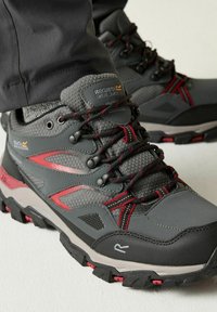 Botas de senderismo exteriores en gris y rojo con suelas texturizadas y cordones negros y rojos, usadas con pantalones oscuros y calcetines grises.