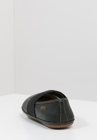 Zapato negro de cuero tipo slip-on con parte superior texturizada y cinta elástica. Presenta una punta redondeada y una suela de goma marrón con costuras en contraste.