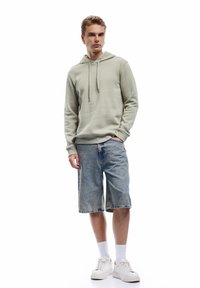 Młody mężczyzna stojący z jedną ręką w kieszeni, ubrany w jasną zieloną bluzę z kapturem, długie dżinsowe szorty, białe skarpetki i białe trampki, na białym tle.