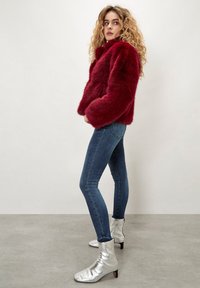 Mujer con cabello rizado rubio que lleva una chaqueta de piel sintética roja, jeans ajustados azules y botas de tobillo plateadas, de pie contra una pared lisa.