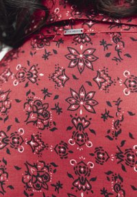 Rode stof met zwart-witte bloemen- en geometrische patronen op de kraag van een kledingstuk met het label "HET IS VROUWEN," met donkere haarstrengen zichtbaar.