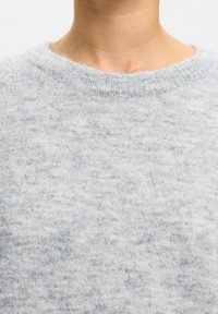 Detailný záber na krk a plecia osoby, ktorá má na sebe mäkký svetlosivý pletený sveter s okrúhlym výstrihom na jednofarebnom pozadí.