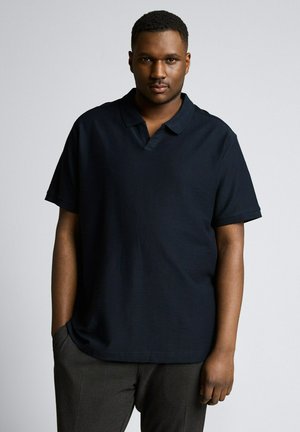 Mann mit kurzen Haaren, der ein dunkelblaues Polohemd und graue Hosen trägt, steht mit einer Hand in der Tasche vor einfarbigem Hintergrund.