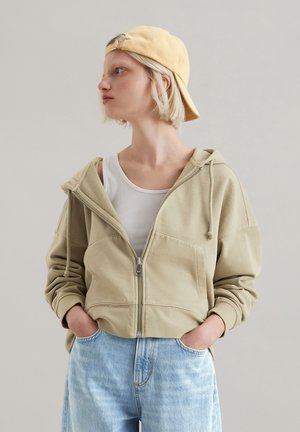 Sweat à capuche beige zipé avec une poche frontale, cordon de serrage, et coupe décontractée, porté sur un débardeur blanc et associé à un jean bleu clair.