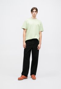 Camiseta de algodón verde claro con mangas cortas y un gráfico en el pecho, combinada con pantalones anchos negros y zapatos naranjas con cordones.