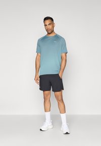Camisa deportiva de manga corta azul clara con un sutil patrón de degradado, combinada con pantalones cortos negros. Zapatillas blancas y calcetines hasta el tobillo completan el conjunto.