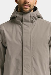 Hellbeige wasserdichte Jacke mit hohem Kragen, Frontreißverschluss und lockerem Schnitt. Verfügt über dezente Markenkennzeichnung an der Seite.