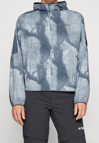 Leichte Jacke mit blau-grauem Muster, Kapuze, Reißverschluss, elastischen Bündchen und Logodetails. Das Material wirkt glatt und wasserabweisend.