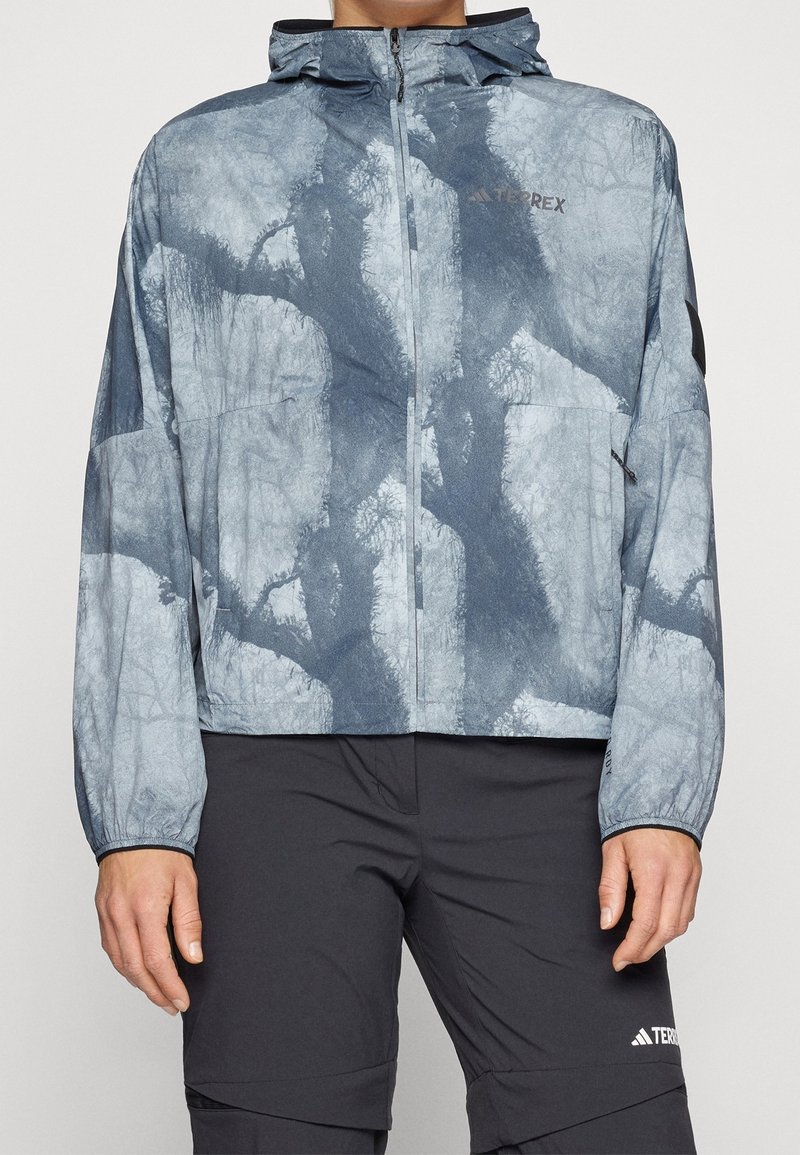 Leichte Jacke mit blau-grauem Muster, Kapuze, Reißverschluss, elastischen Bündchen und Logodetails. Das Material wirkt glatt und wasserabweisend.