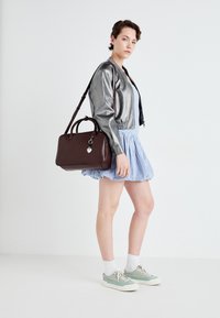 Brown leather handbag with a top handle and crossbody strap, a silver charm, worn with a metallic bomber jacket and a blue checkered skirt.