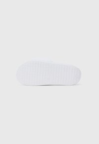 Vit sneakersula med en texturerad, mönstrad greppyta och en cirkulär logotyp i mitten. Platt design, tillverkad av gummi.