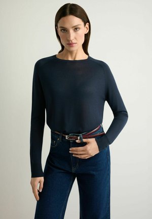 Kobieta w dopasowanym granatowym swetrze wpuszczonym w ciemnoniebieskie, wysokowcześniowe dżinsy z brązowym paskiem, stojąca na tle jednolitego jasnego tła.