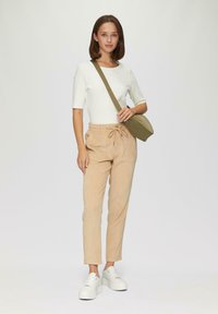 Beige Cordhosen mit elastischem Bund und Kordelzug, kombiniert mit einem taillierten weißen Rippenshirt und weißen Plateauschuhen. Olivgrüne Umhängetasche.