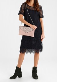 Musta pitsimekko lyhyillä hihoilla ja kaarevalla helmalla, yhdistettynä vaaleanpunaiseen kirjepussiin ja vihreisiin nilkkasaappaisiin.