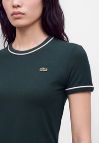 Femme portant un t-shirt à manches courtes vert foncé avec une bordure claire au niveau du col et des manches et un petit logo crocodile sur la poitrine.