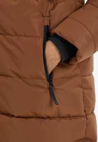 Bruine pufferjack met horizontale stiksels, voorzien van een ritsvak met een zwarte rits en een aansluitende zwarte manchet aan de mouw.
