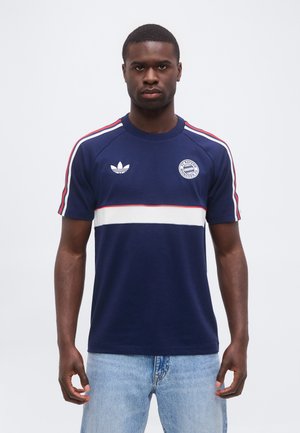 Uomo che indossa una maglietta a maniche corte Adidas FC Bayern Monaco blu navy con strisce rosse e bianche, abbinata a jeans azzurri chiari.
