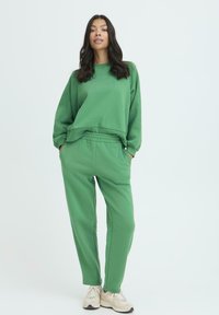 Zeleni pamučni dukserica s manžetama, uparena s odgovarajućim zelenim pamučnim trenirkama koje imaju elastičnu sredinu i bočne džepove.