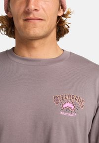 Szara bluzka z długim rękawem i okrągłym dekoltem, z kolorową grafiką "Billabong Australia" na lewej stronie klatki piersiowej.