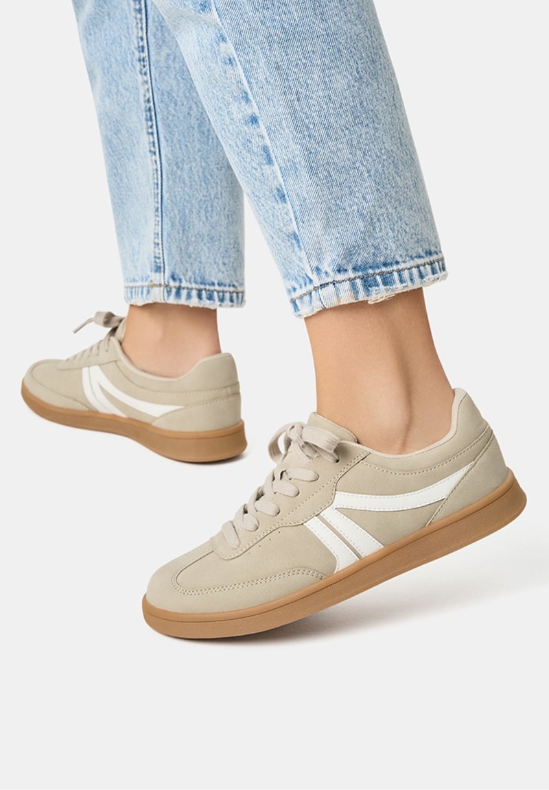 Baskets beiges en daim et caoutchouc, avec des rayures blanches, un bout rond et une semelle en caoutchouc marron. Portées avec un jean court bleu clair.