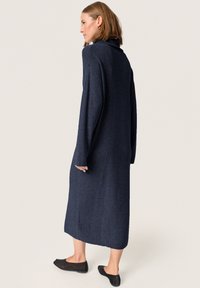 Rochie tricotată, de culoare bleumarin, cu mâneci lungi și guler înalt. Are un croi lejer, până la genunchi, și o textură cu nervuri. Poartă balerini negri.
