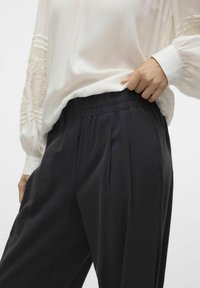 Zwarte hoge taille broek met plooien en een elastische tailleband. Draagt een witte blouse met textuuraccenten op de mouwen.
