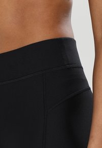 Czarne legginsy o wysokim stanie z gładkiego, elastycznego materiału, z płaskimi szwami i eleganckim designem. Zbliżenie na obszar pasa.