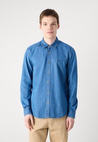 Camicia in denim azzurro chiaro con chiusura a bottoni, colletto classico e maniche lunghe. Caratterizzata da bottoni dorati e un piccolo logo nero sul petto.