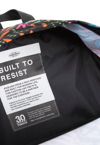 Mochila con un exterior colorido y estampado que presenta acentos en negro, naranja y azul. En el interior, un forro negro suave y una etiqueta de cuidado.
