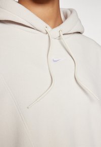 Close-up van een persoon die een off-white hoodie met koordjes draagt en een klein geborduurd Nike-logo op de borst heeft.