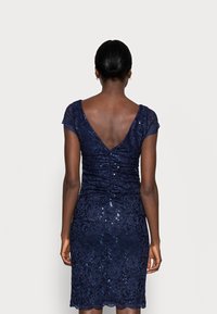 Femme à la peau foncée et aux cheveux en chignon bas, portant une robe en dentelle bleu marine ajustée avec des sequins et un dos en V profond, se tenant contre un fond uni.