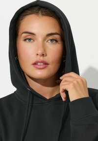 Hoodie nero con tasca frontale, cordini sul cappuccio e polsini a costine. La texture appare morbida e liscia, con una vestibilità rilassata.