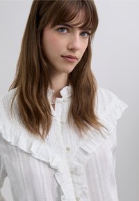 Vit skjorta med knappar fram, volangkrage och texturerat tyg. Har tunna vertikala ränder och blanka knappar.