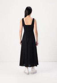 Sort maxi kjole med firkantet ryg og smocket barm, der har brede stropper. Bæres med hvide høj-hælede sneakers og tekstureret stof.