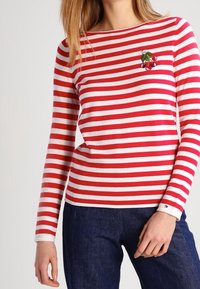 Rød og hvidstribet langærmet skjorte med rund hals og broderet kirsebærdesign på brystet, parret med mørke denimjeans.