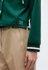 Κοντινό πλάνο ενός ατόμου που φοράει μπεζ παντελόνι και πράσινο αθλητικό μπουφάν Marc Cain Sports με λευκή ρίγα στο μανίκι και λογότυπο κοντά στην τσέπη.