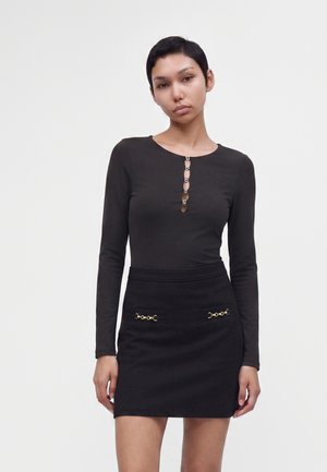 Jeune femme aux cheveux courts portant un haut noir à manches longues et une jupe taille haute assortie avec des détails dorés sur les poches.