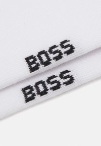 Chaussettes en coton blanches avec le logo "BOSS" noir brodé, présentant une texture côtelée et un design sans couture.