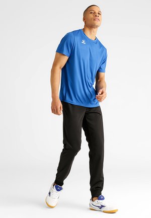T-shirt atletico blu traspirante, pantaloni neri da atleta e sneakers bianche con dettagli blu. Il modello si trova in una posa dinamica.