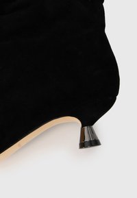Scarpa nera in suede con tacco alto, punta rotonda e un accento metallico distintivo sul tacco. Texture liscia con una finitura opaca.