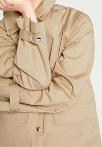 Personne portant une veste beige à boutons-pression, bras croisés, une main reposant près du visage montrant un petit tatouage "USA".