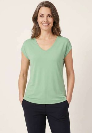 Femme souriante aux cheveux bruns portant un t-shirt vert clair à manches courtes et col en V ainsi qu’un pantalon sombre, mains dans les poches, sur un fond clair uni.