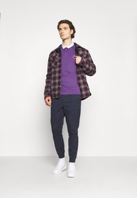 Junger Mann, der ein lila Poloshirt, eine karierte Jacke, navy Jogginghosen und weiße Sneakers trägt, steht vor einem schlichten weißen Hintergrund.