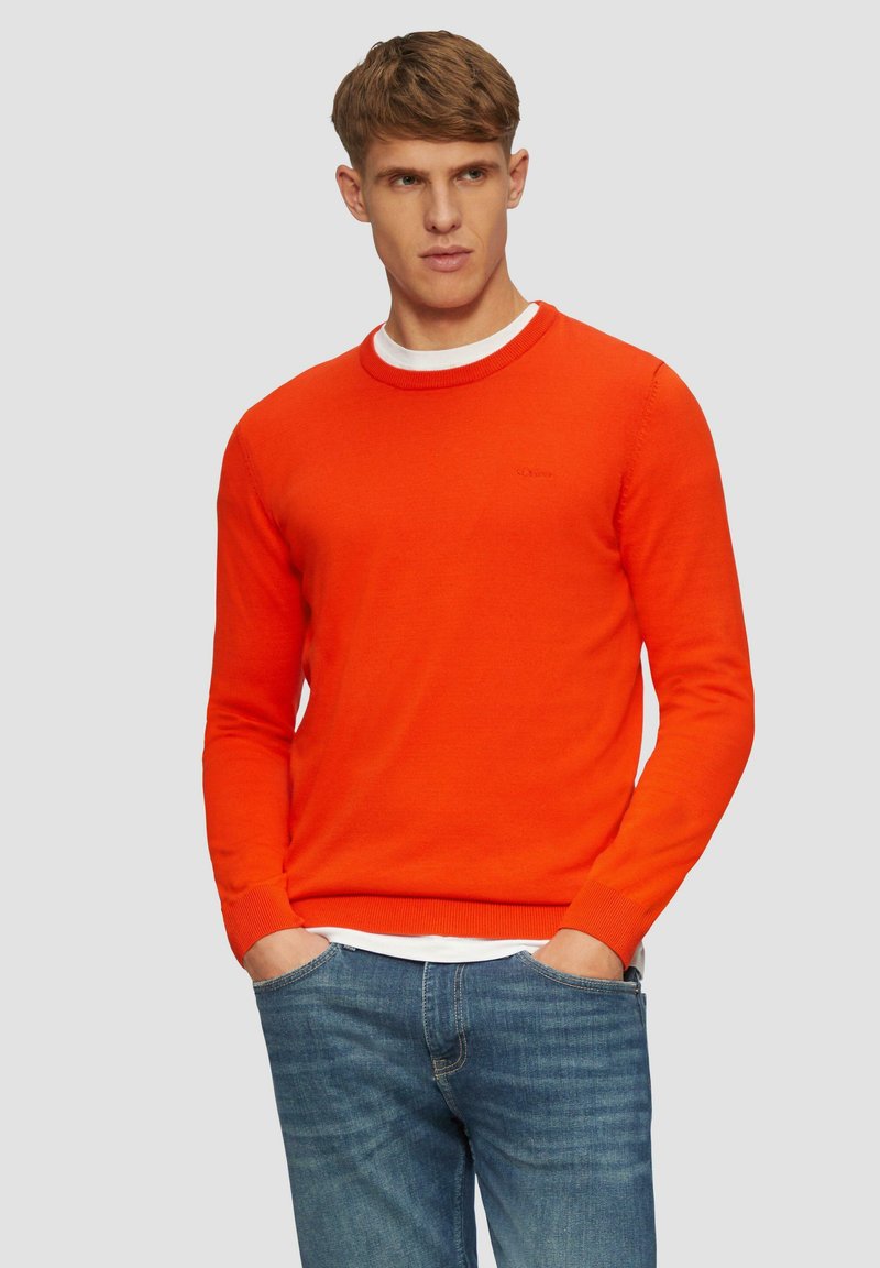 Orange Crewneck-Pullover mit gerippten Bündchen und Saum. Über einem weißen Hemd getragen, kombiniert mit blauen Jeans. Glatte Textur, minimale Markenkennzeichnung.