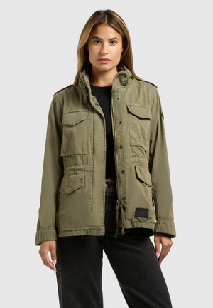 Nő olívazöld, több zsebbel ellátott munkaruhakabátban, fekete felső és fekete nadrág fölött, egyszínű háttér előtt áll.