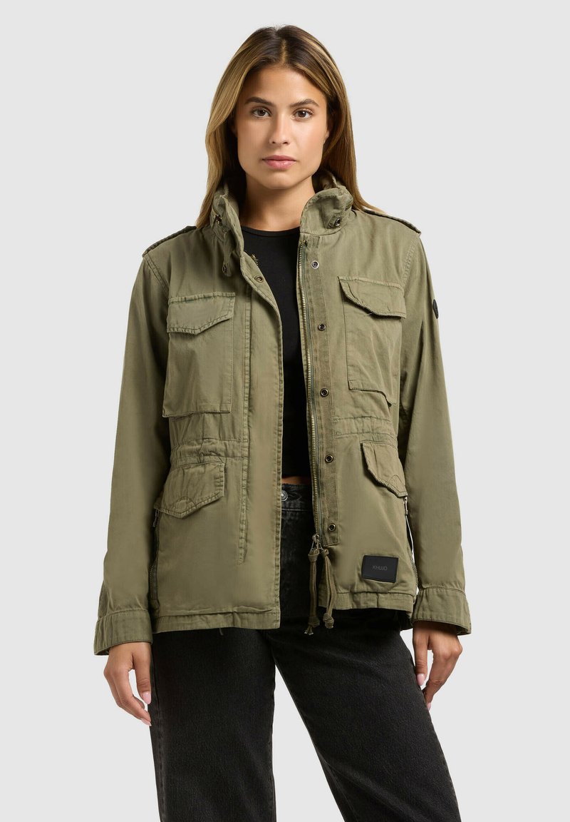 Mujer con chaqueta utilitaria verde oliva con múltiples bolsillos delanteros, encima de una camiseta negra y pantalones negros, posando frente a un fondo liso.