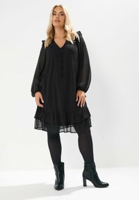 Schwarzes, geschichtetes Kleid mit einem strukturierten Pünktchenmuster, V-Ausschnitt und Rüschensäumen. Getragen mit schwarzen Knöchelstiefeln und blickdichten Strumpfhosen.