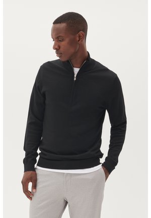 Svart zip-up sweatshirt med långa ärmar, tillverkad av mjukt tyg. Har en hög krage och ribbade manschetter och nederkant. Bärs över en vit skjorta.